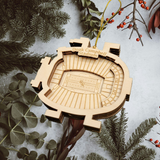A wooden ornament resembling a football stadium is hanging from a string, surrounded by pine branches and greenery.