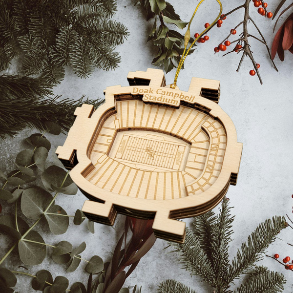 A wooden ornament resembling a football stadium is hanging from a string, surrounded by pine branches and greenery.