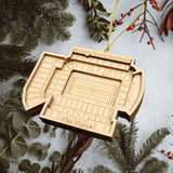 A wooden ornament resembling a stadium is hanging from a string, surrounded by pine branches and greenery.