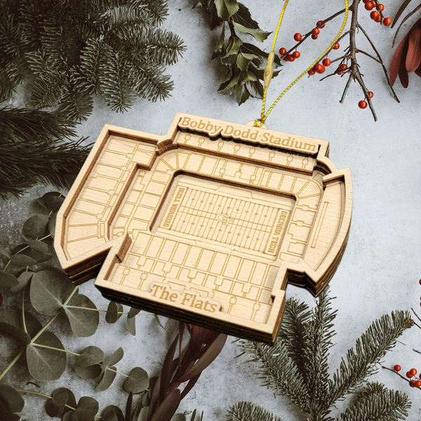A wooden ornament resembling a stadium is hanging from a string, surrounded by pine branches and greenery.