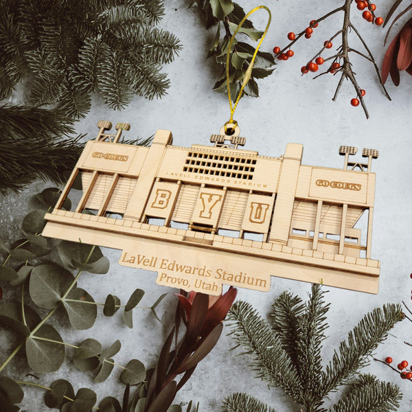A wooden ornament depicting LaVell Edwards Stadium in Provo, Utah, hanging among pine branches and greenery.