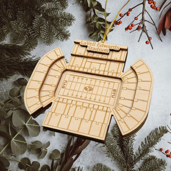 A wooden ornament resembling a football stadium is displayed among pine branches and greenery, with the text "LaVell Edwards Stadium" visible.